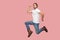 Portrait of carefree bearded hipster young man in white shirt and blue jeans with hat jumping in super mario pose and looking at