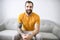 portrait of casual young man with beard and tattoo and Burnt Orange shirt sit on the sofa at home