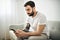 portrait of casual young man with beard and tattoo and white shirt sit on the sofa at home with cellphone