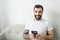 portrait of casual young man with beard and tattoo and white shirt sit on the sofa at home with cellphone