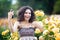 A portrait of Caucasian woman with dark curly hair taking selfie near yellow rose bushes in a rose garden, video call, chat, smile