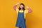 Portrait of charming bright girl teenager in denim sundress keeping hands on french beret isolated on yellow wall