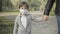 Portrait of charming brunette little boy in Covid-19 face mask walking in park holding hand of unrecognizable father