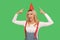 Portrait of charming joyful adult woman in stylish overalls pointing at party cone on her head, celebrating birthday
