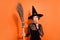Portrait of charming witch person finger touch chin pouted lips look empty space hold broom  on orange color