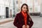 Portrait charming young woman in elegant burgundy coat with fashionable orange scarf with trendy hairstyle on street in city.