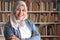 Portrait of cheerful Asian muslim female librarian wearing hijab and smiling, woman standing against books in library