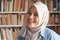 Portrait of cheerful Asian muslim female librarian wearing hijab and smiling, woman standing against books in library