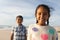 Portrait of cheerful biracial girl standing with brother at beach against sky