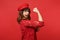 Portrait of cheerful strong young woman in lace dress and cap showing biceps, muscles isolated on bright red wall