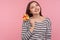 Portrait of cheerful woman in striped sweatshirt holding sweet round rainbow candy lollipop, pink background