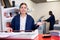 Portrait of cheerful young female typographer in uniform using paper cutter during work in printer shop