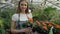 Portrait of cheerful young woman garden worker in apron smiling and holding flower in hands in greenhouse