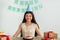 Portrait of cheery Indian teen girl in party hat sitting at table with gift boxes and birthday cake with candles at home