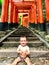 Portrait of a cherful little baby with red colorful toriis of Fushimi Inari Taisha shrine on background, Kyoto, Japan