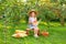 Portrait of child in apple orchard. Little girl in straw hat and dress, sits on bench holding wicker basket with apples