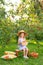 Portrait of child in apple orchard. Little girl in straw hat, striped dress, sits on bench holding basket with apples