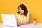 Portrait of concentrated woman manager sitting at table with laptop on yellow background in studio, lady looks astonished on