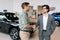 Portrait of confident car dealer in business suit welcoming young man customer in showroom. Professional salesman