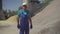 Portrait of confident Caucasian man in blue uniform and safety helmet walking on manufacturing site along piles of bulk