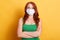 Portrait of confident healthy wavy red haired girl, wearing medical mask, folded arms, stays isolated over yellow background,