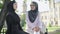 Portrait of confident positive woman in hijab and eyeglasses sitting with friend outdoors and talking. Young beautiful