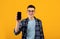 Portrait of cool young guy showing smartphone with empty screen on orange studio background, mockup for design