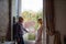 Portrait of Couple with Age Difference Standing near Opened Window inside the House During Summer Sunny Day.