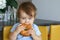 Portrait of cute baby boy with stylish haircut holding and eating big bagel sitting on yellow chair at home kitchen