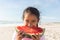 Portrait of cute biracial girl eating large fresh watermelon slice at beach on sunny day