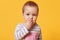 Portrait of a cute brooding girl with short fair hair keeping fingers in her mouth. A serious kid stands in front of camera