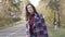 Portrait of cute Caucasian brunette girl hitchhiking on small road in the forest. Positive young woman in plaid jacket