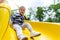 Portrait of cute little cute caucasain blond toddler boy enjoy having fun sliding down modern plastic slide at outdoors