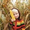 Portrait of cute playful smiling child in colorful knitted sweater playing with fresh corn cob on autumn cornfield. Childhood,