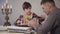 Portrait of cute smart Caucasian boy placing chess on board and talking to father sitting at the foreground and using