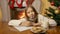 Portrait of cute smiling girl sitting behind table at living room and writing letter to Santa Claus