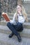 Portrait of Cute Thinking and Tranquil Caucasian Blond Female With Book Sitting Straight on Stairs Outdoors and Reading.