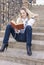 Portrait of Cute and Tranquil Caucasian Blond Woman Reading Book While Sitting Straight on Stairs Outdoors