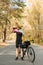 Portrait of cyclist in sportswear standing with bicycle on road in autumn forest and drinking water from bottle. Thirsty cyclist