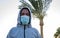 Portrait of a dark-skinned man in a medical mask and hoodie against the sky and palm trees. Concept:street safe distance, coronavi