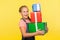 Portrait of delighted little girl with braid in denim overalls holding lot of gift boxes and smiling to camera