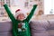 Portrait dreaming boy with closed eyes in Santa hat and Christmas costume sitting at home hands up. New Year Holidays