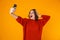 Portrait of ecstatic woman holding and taking selfie photo on cell phone while standing isolated over yellow background