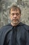 Portrait of an elderly monk 45-50 years old with a beard in a black robe on the background of a textured wall, looking directly in