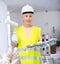 Portrait of elderly positive woman painter in protective helmet and yellow vest who paints walls with paint roller