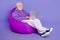 Portrait of elderly retired trendy cheery man pensioner sitting in bag writing email isolated over bright violet purple