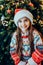 Portrait of emotional happy smiling little girl in Santa hat and festive knitted sweater standing near the Christmas tree.