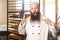 Portrait of excited adult businessman baker with beard in white uniform standing in bakery and waiting for online order by phone,