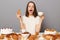 Portrait of excited amazed woman with brown hair sitting at table isolated over gray background, breaking diet, drinking coffee or