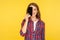 Portrait of excited cheerful girl in shirt covering half head with mobile phone, hiding half face with cellphone and smiling at